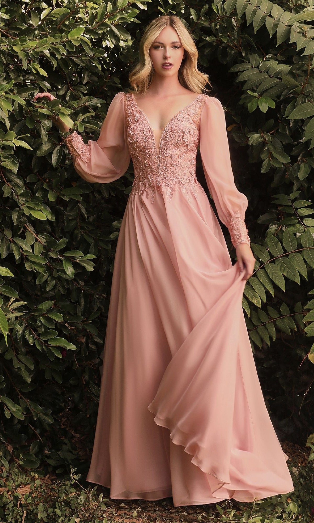 A woman in the Modest Long Sleeve Formal Gown: Ladivine CD0183, featuring lace details and sheer sleeves, stands outdoors amid lush greenery—a stunning choice for a mother of the bride.